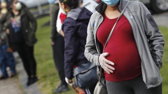 A pregnant woman wearing a face mask and gloves holds her belly.