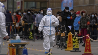 A health worker wearing a protective suit walks by masked residents who wait in line to get their throat swab