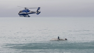 A helicopter flies overs an upturned boat with a survivor sitting on the hull.