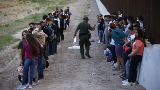 A group of migrants stand next to the border wall as a Border Patrol agent takes a head count.