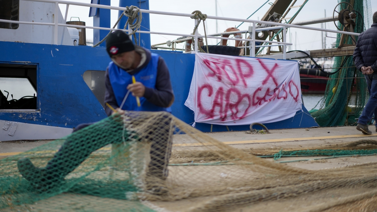 A fisherman mends a net in front of his fishing boat with a banner reading "stop for gasoline increase"