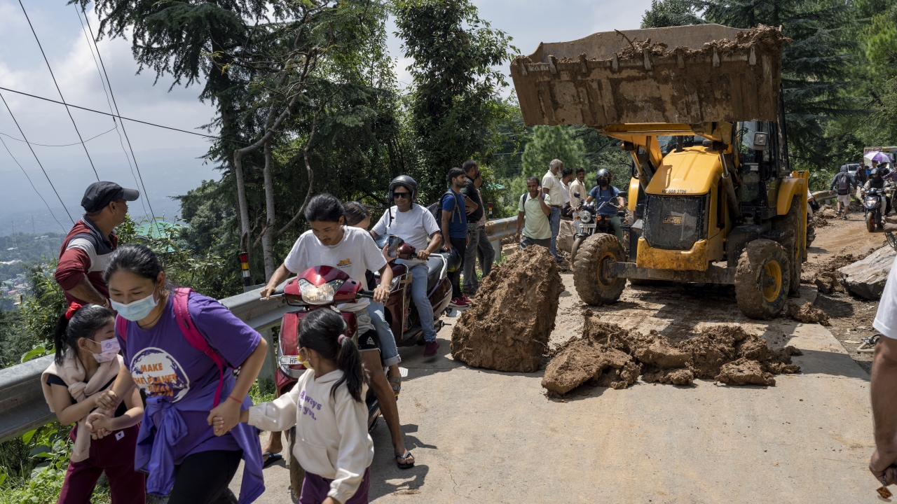 People rush past an earthmover clearing a road of a big rock that came down with mud and plant debris following monsoon rains